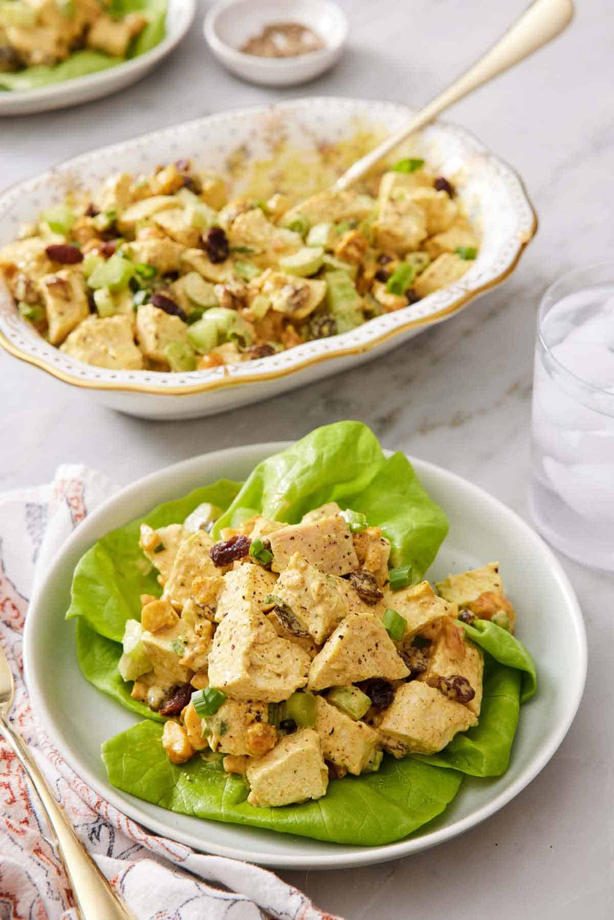 A plate of curried chicken salad over lettuce with a platter of more chicken salad in the background.
