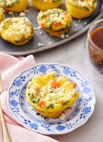 A plate with an egg muffin topped with crumbled feta cheese. A platter with more egg muffins and a coffee in the background.