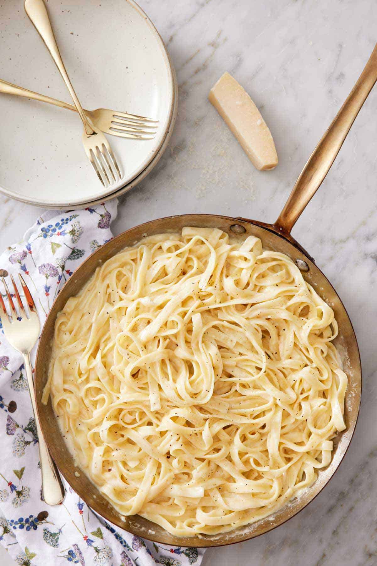 Overhead view of a skillet of fettucine alfredo with a stack of plates, fork, and parmesan beside it.