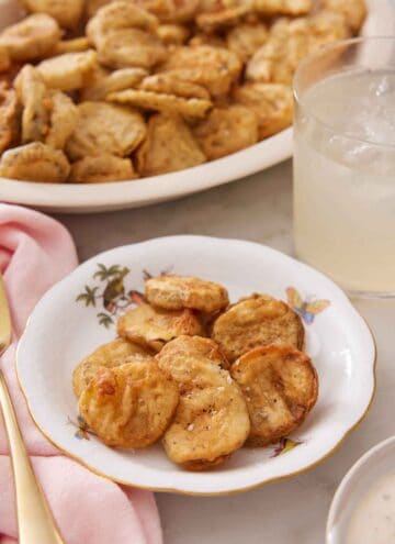 A plate of fried pickles with a drink and platter of more fried pickles in the background.