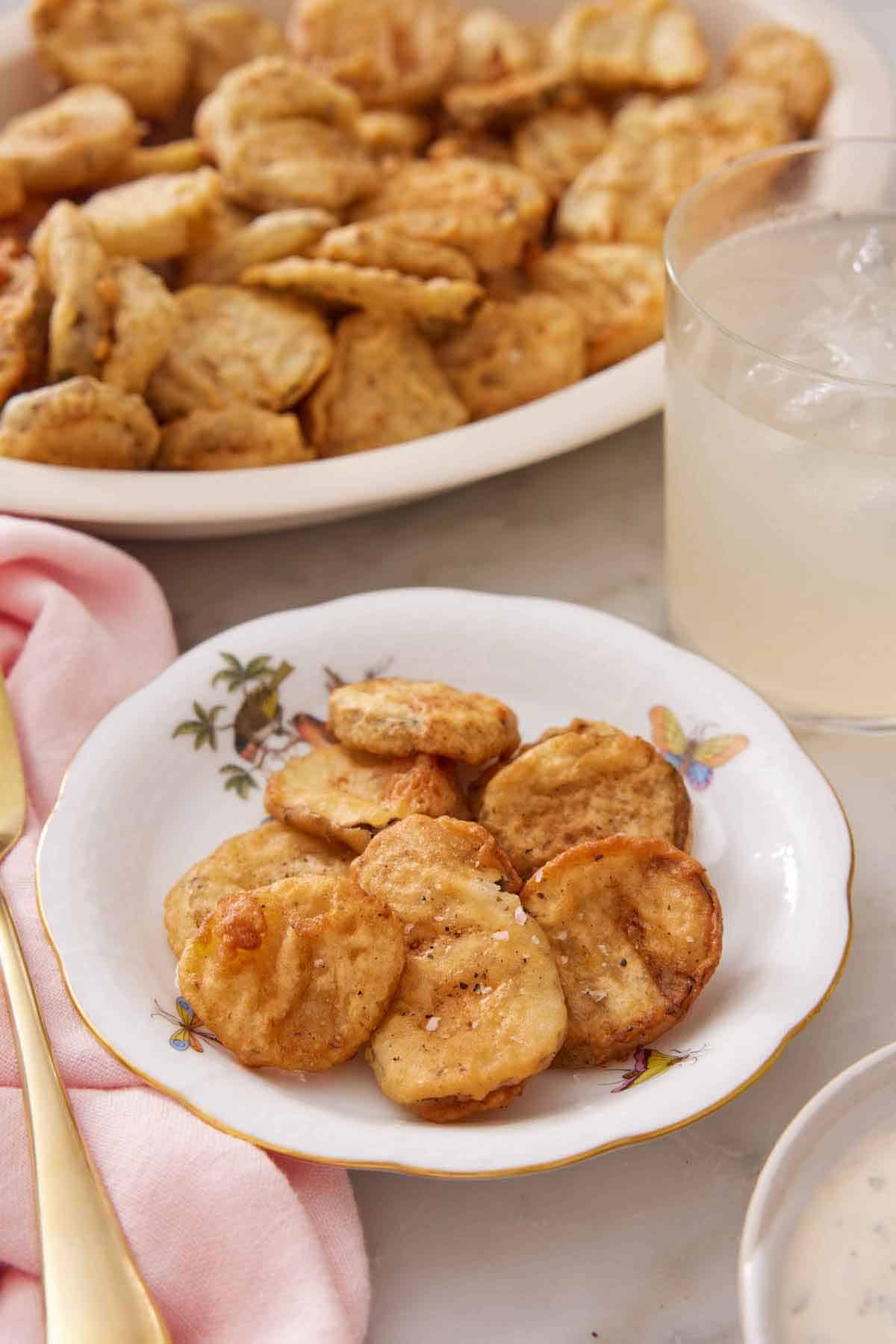 A plate of fried pickles with a drink and platter of more fried pickles in the background.