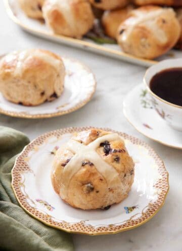 A plate with a hot cross bun with a second plate in the back along with a platter and a cup of coffee.