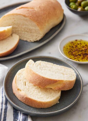 A plate with two slices of Italian bread with the rest of the cut loaf in the background along with a plate of seasoned oil.