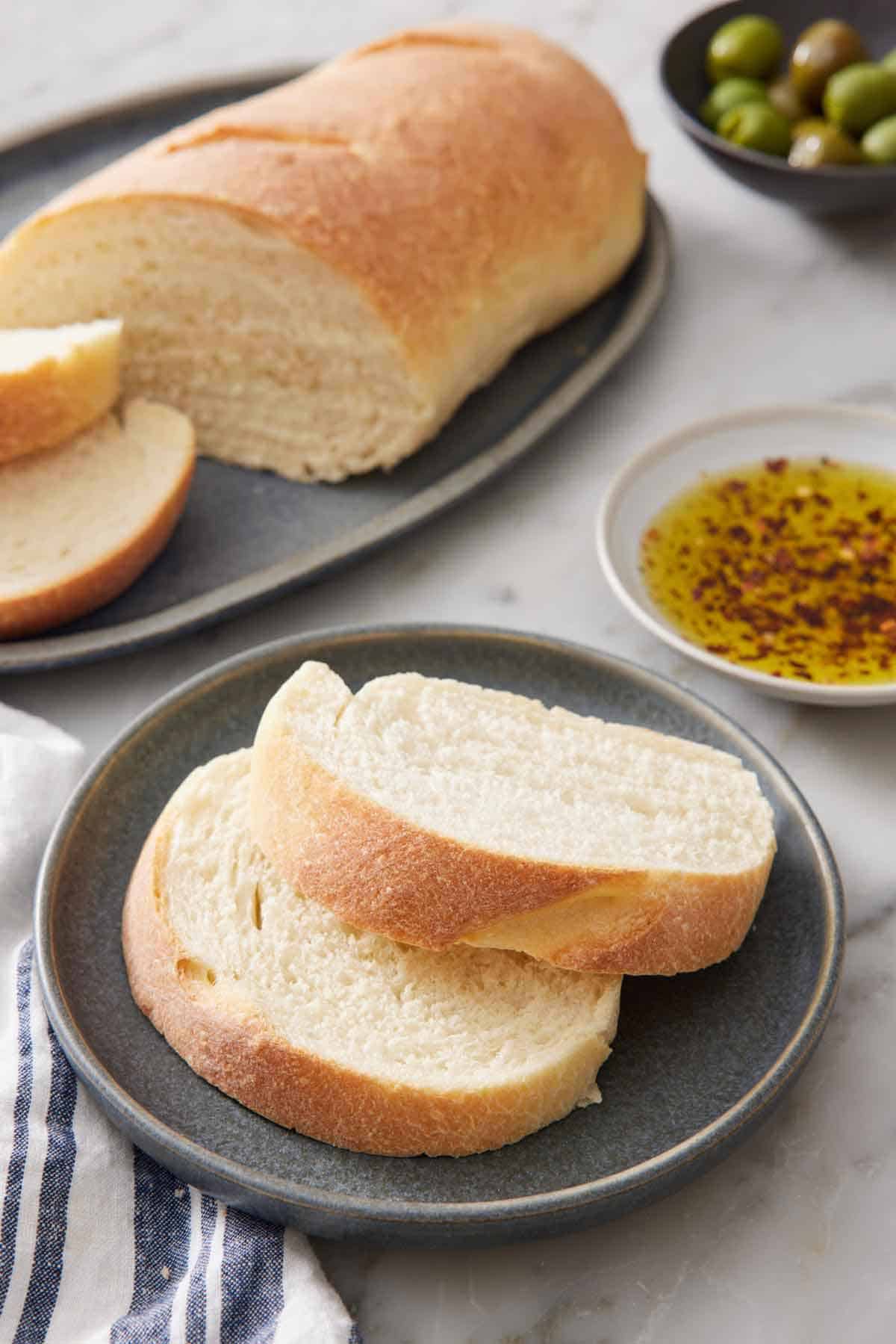 A plate with two slices of Italian bread with the rest of the cut loaf in the background along with a plate of seasoned oil.