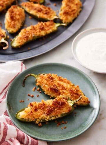 A plate with two jalapeno poppers with a bowl of dipping sauce on the side. A platter with more jalapeno poppers in the background.