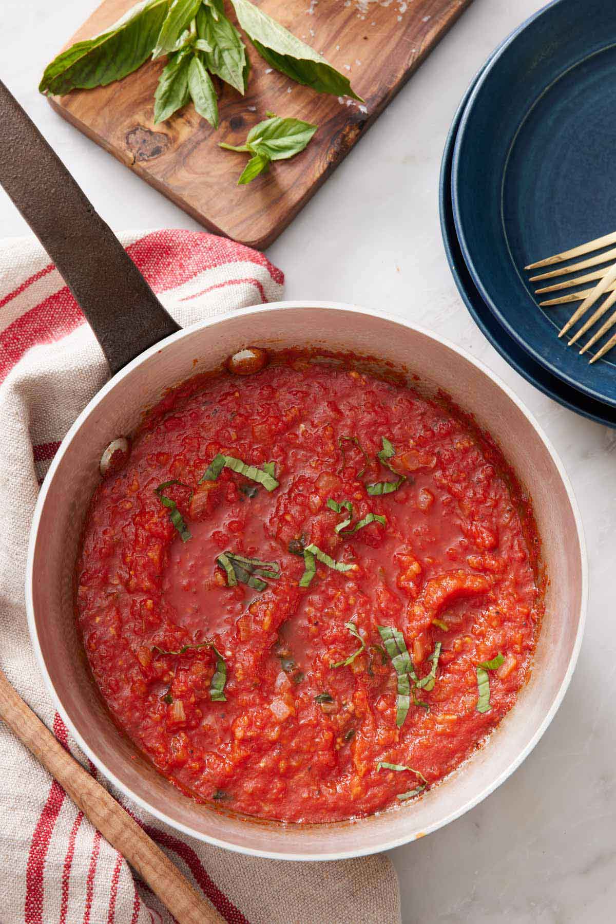 An overhead view of a pot of marinara sauce with some fresh ribboned basil as garnish and some leaves off to the side along with plates and forks.