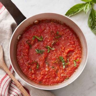 An overhead view of a pot of marinara sauce with a wooden spoon and fresh basil on the side.
