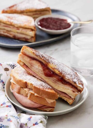 A plate with a Monte Cristo sandwich cut in half with another one in the background along with a glass of water.