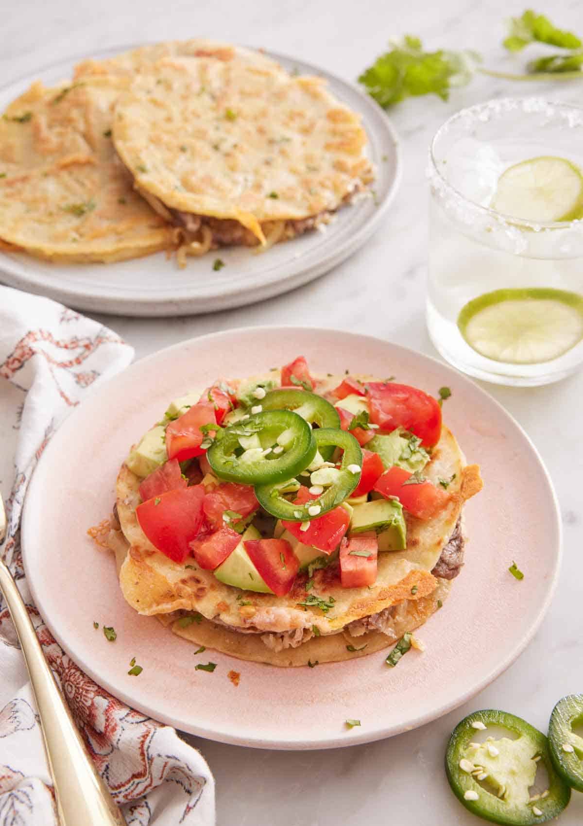 A plate of mulita topped with tomatoes, avocados, and jalapenos. A drink in the back and more mulitas on a plate.