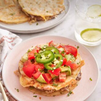 Pinterest graphic of a plate of mulita topped with tomatoes, avocados, and jalapenos. A drink in the back and more mulitas on a plate.
