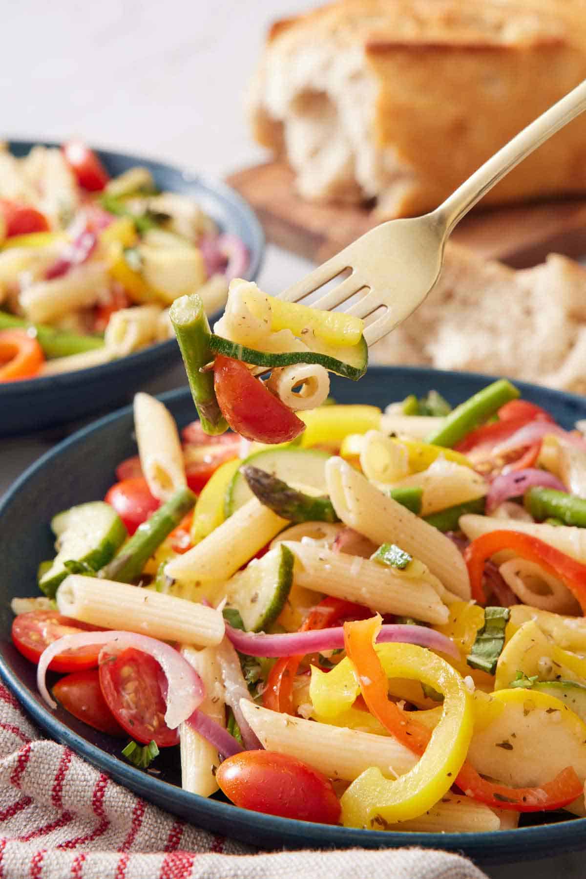 A fork lifting up a bite of pasta primavera from a bowl. Torn bread in the background.