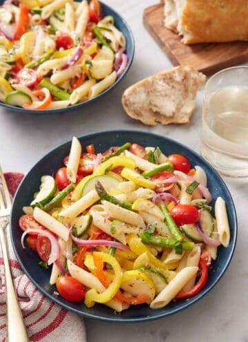 A bowl of pasta primavera with a second bowl in the background and bread torn on the side with a glass of wine.