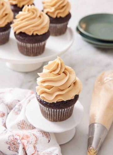A cupcake with peanut butter frosting on top with more frosted cupcakes in the background. A piping bag and plates off to the side.