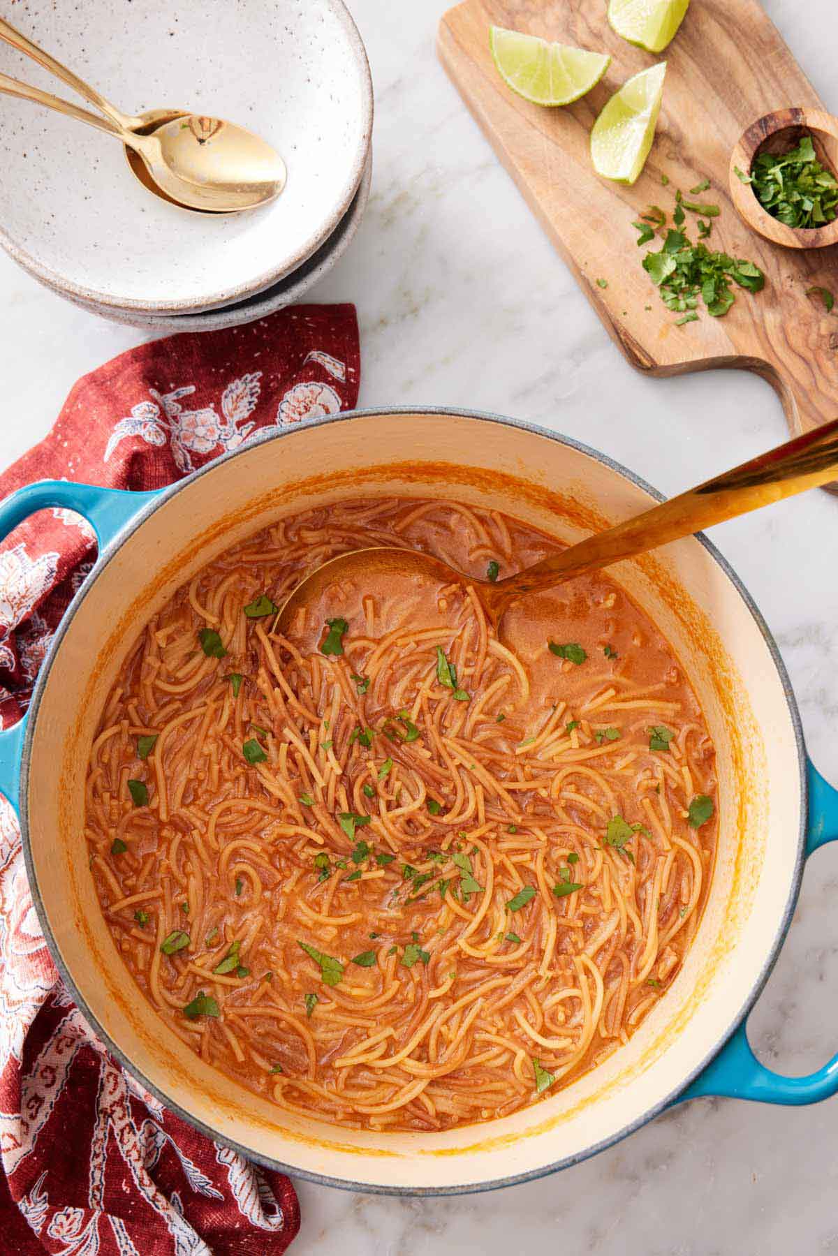 Overhead view of a dutch oven with sopa de fideo with a ladle inside. A board with chopped garnish on the side with some bowls and spoons.