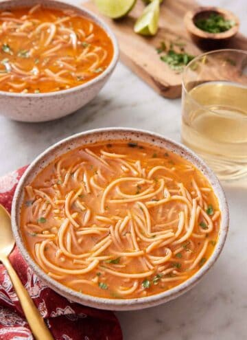 A bowl of sopa de fideo with a drink and second bowl in the background with some garnish on the side.