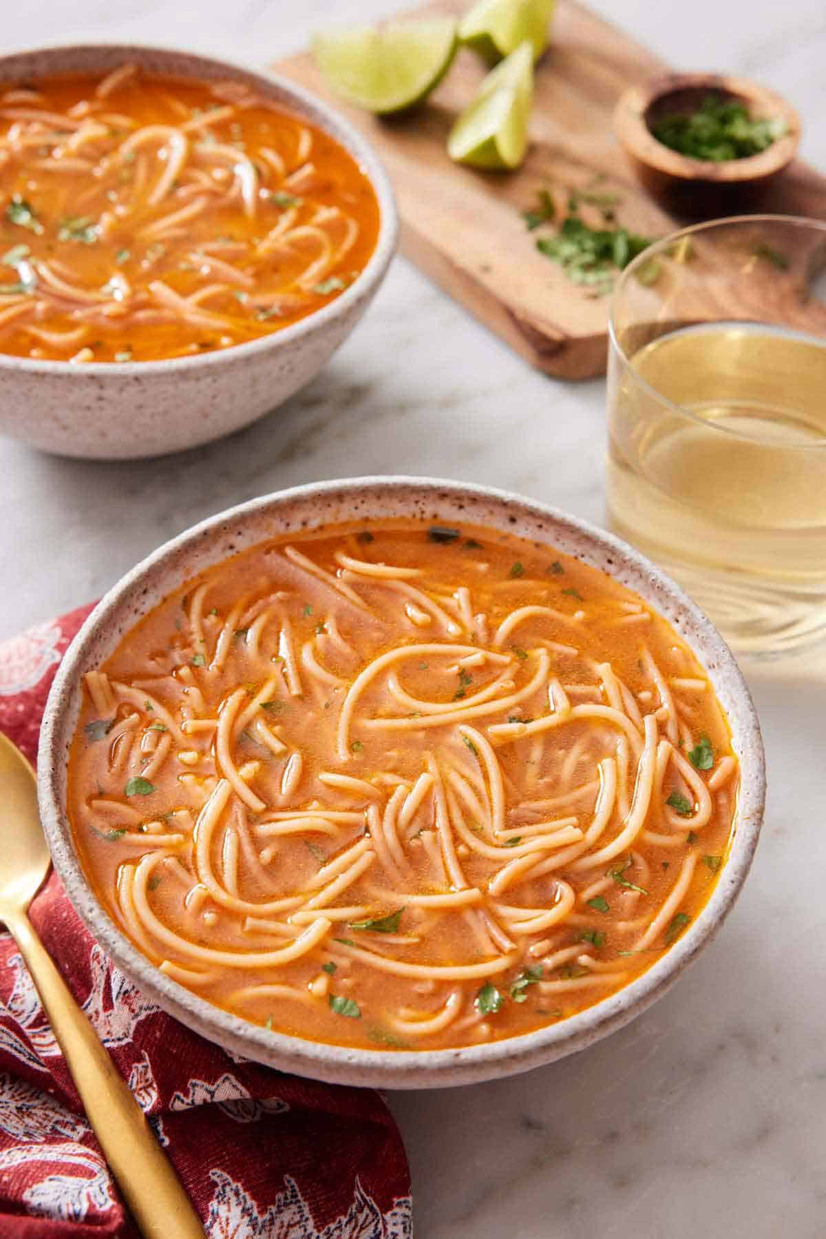 A bowl of sopa de fideo with a drink and second bowl in the background with some garnish on the side.