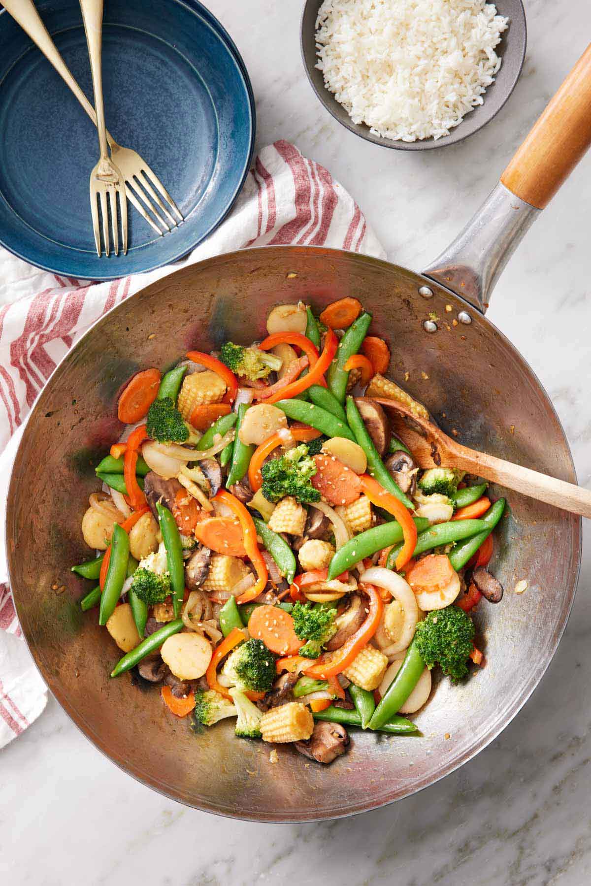 Overhead view of a wok of vegetable stir fry. Plates, forks, and rice off to the side.