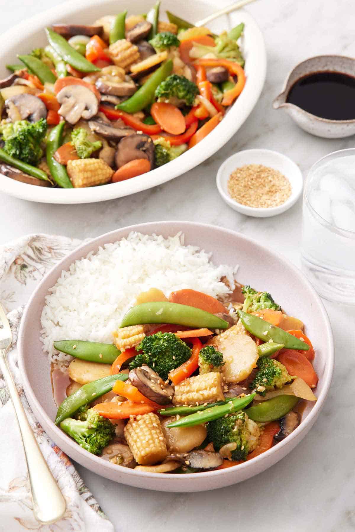 A plate of vegetable stir fry with rice. A platter with more vegetable stir fry in the background along with a bowl of sesame seeds and sauce.