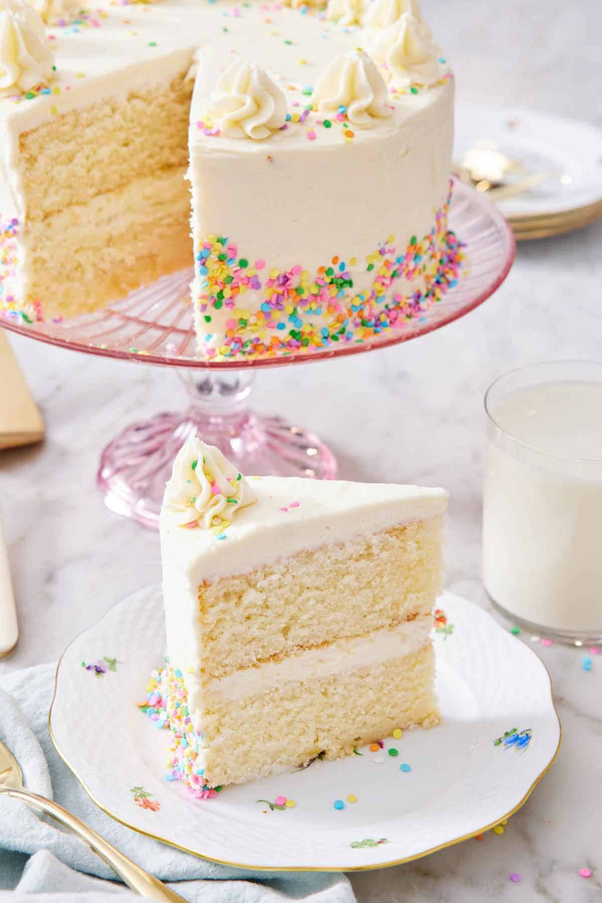 A slice of white cake on a plate with the rest of the cake in the background on a cake stand.