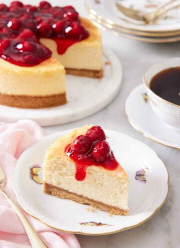 A slice of air fryer cheesecake on a plate with cherry sauce on top. The rest of the cheesecake in the background along with a mug of coffee.