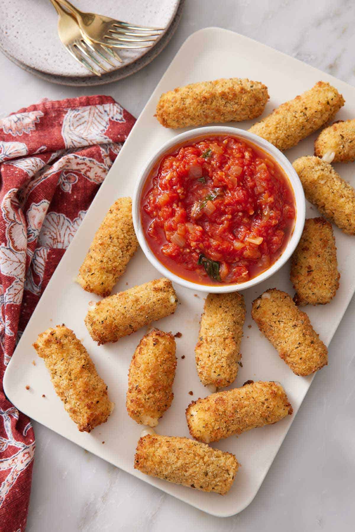 Overhead view of a platter of air fryer mozzarella sticks with a bowl of marinara sauce. Plates and forks on the side.