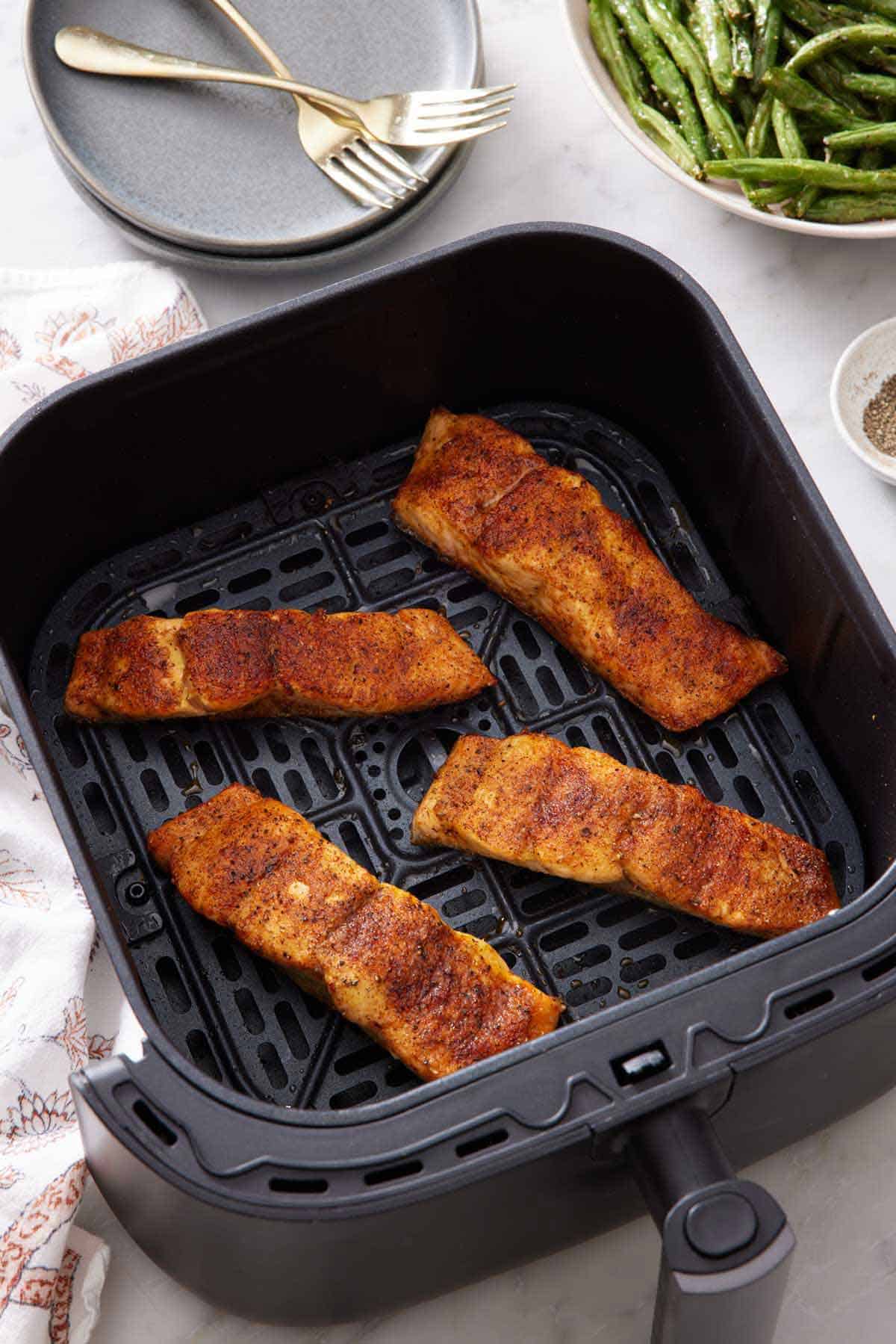 Four pieces of salmon in an air fryer basket. Green beans on the side along with a stack of plates with forks.