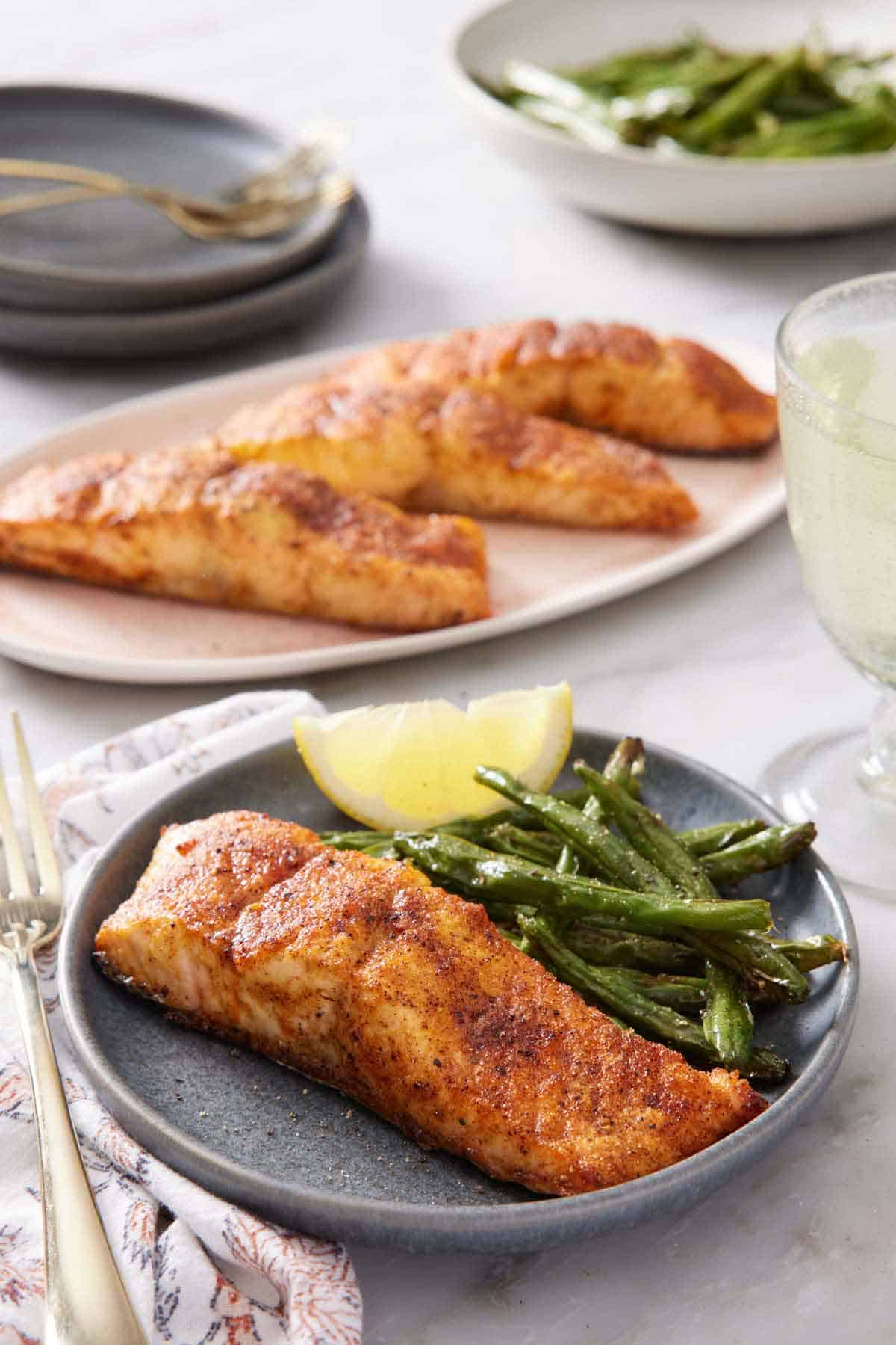 A plate with air fryer salmon along with green beans and a lemon wedge. A platter of more salmon in the background and plate of green beans.