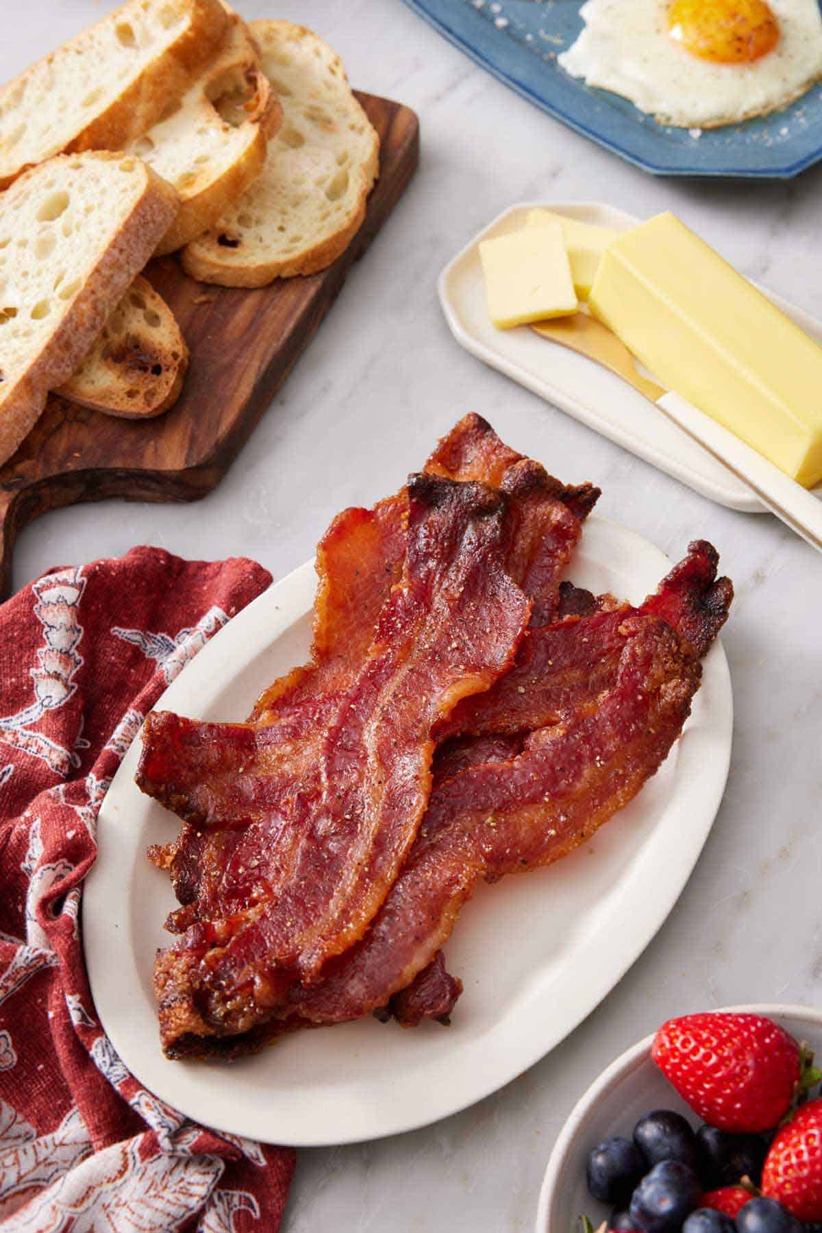 A plate of candied bacon with butter, bread, berries, and eggs around it.