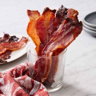 A glass with candied bacon. A platter in the back with more candied bacon and a stack of plates.