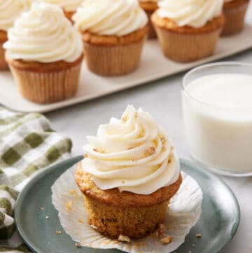 A plate with a carrot cake cupcake with a glass of milk and platter of more cupcakes in the background.