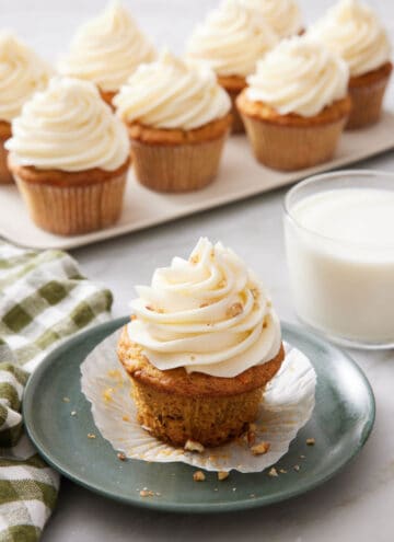 A plate with a carrot cake cupcake with a glass of milk and platter of more cupcakes in the background.