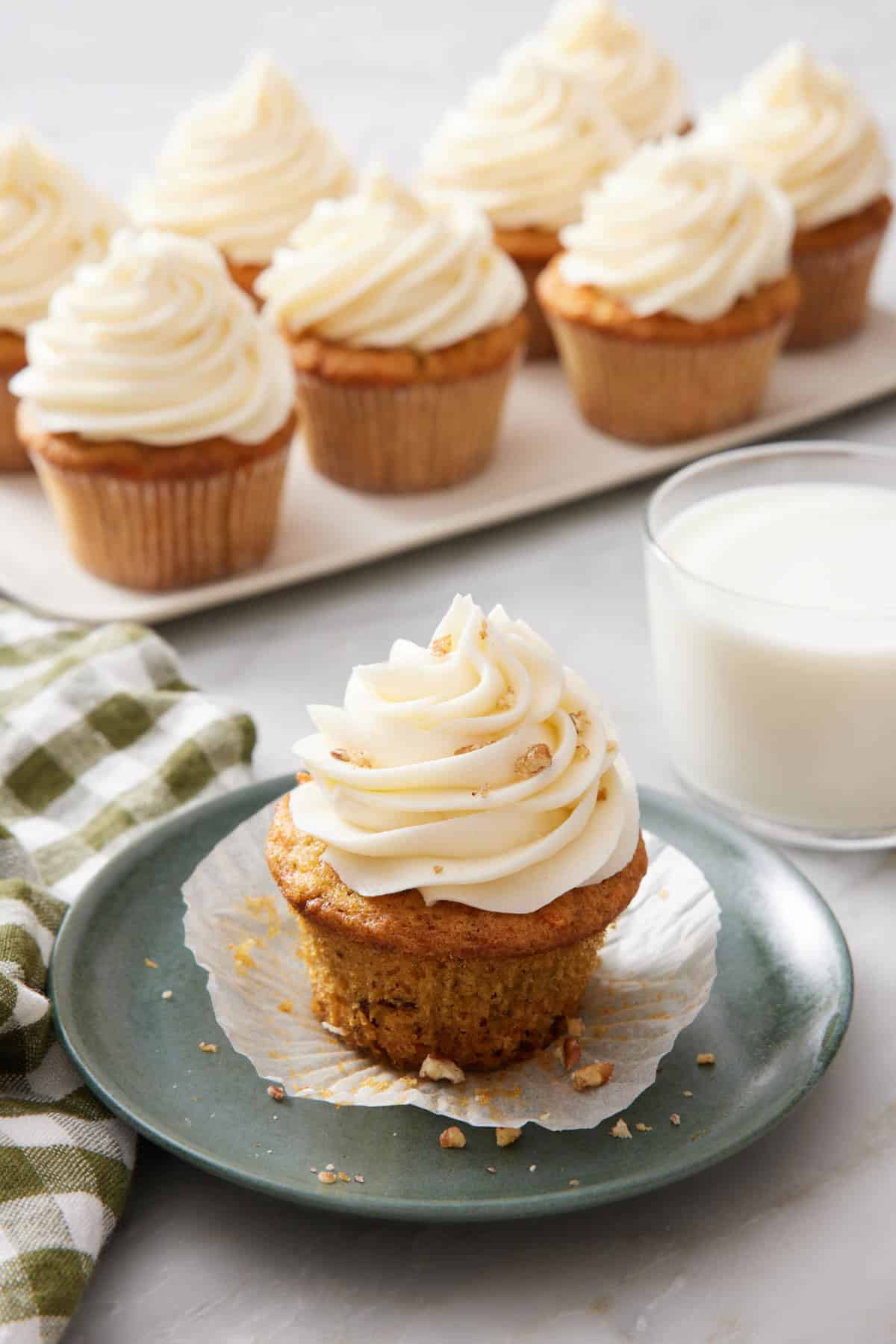 A plate with a carrot cake cupcake with a glass of milk and platter of more cupcakes in the background.