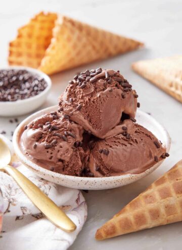 A plate with three scoops of chocolate ice cream topped with chocolate sprinkles. Waffle cones scattered around and a bowl of sprinkles in the back.