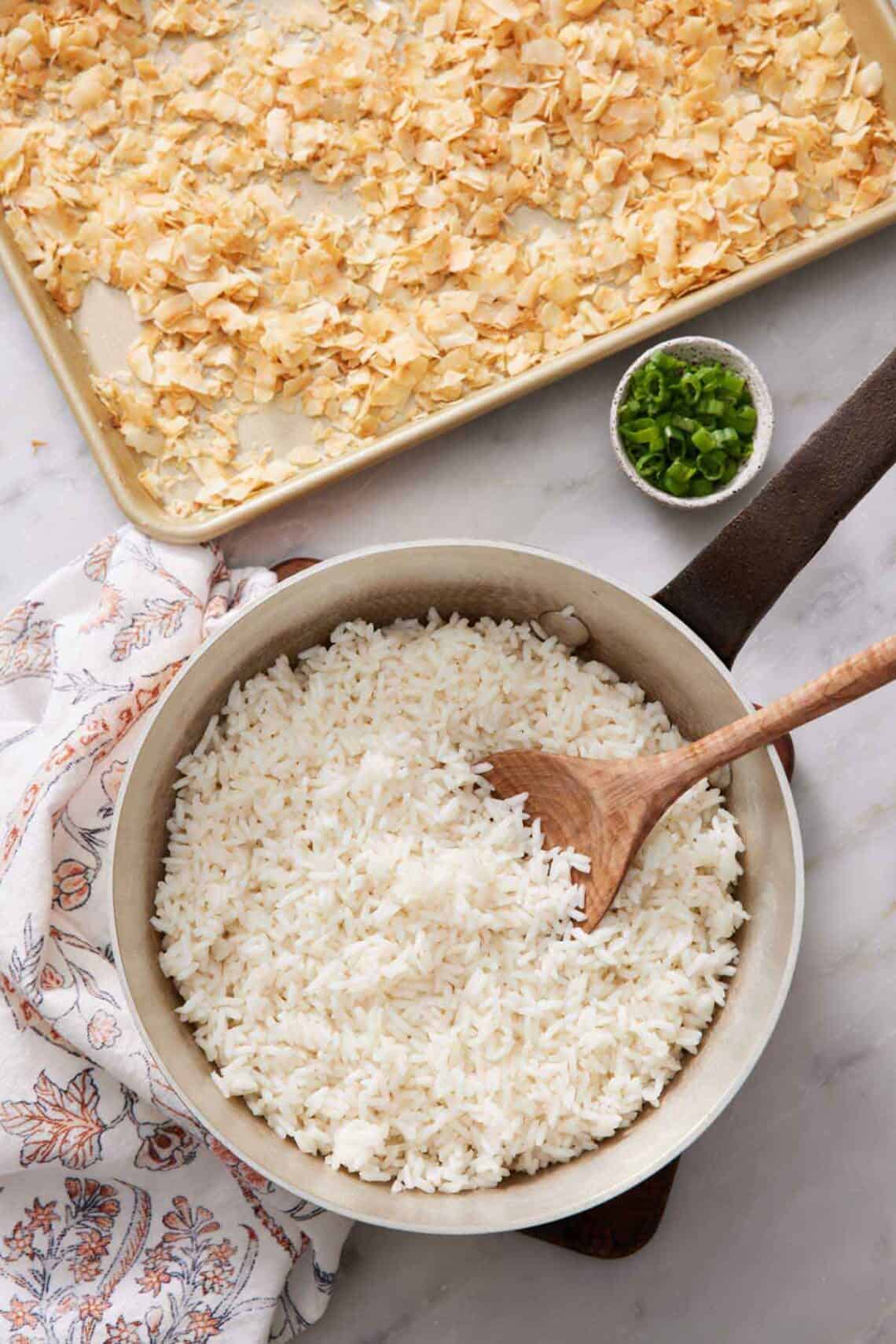An overhead view of a pot of coconut rice with a spoon. A sheet pan of toasted coconut and bowl of green onions beside it.