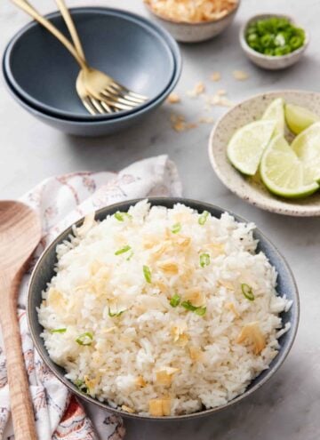 A bowl of coconut rice topped with green onions and toasted coconut. Bowl of limes in the back along with more garnishes and bowls with forks.
