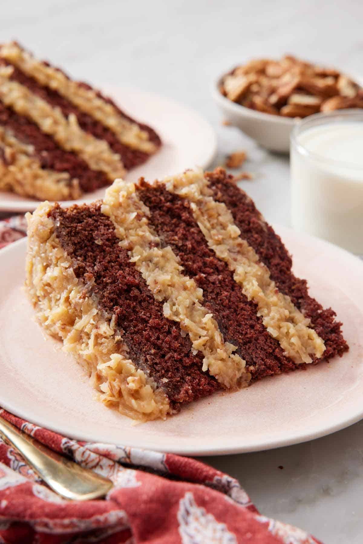 A plate with a slice of German chocolate cake with a glass of milk beside it. A second cake and a bowl of pecans in the background.