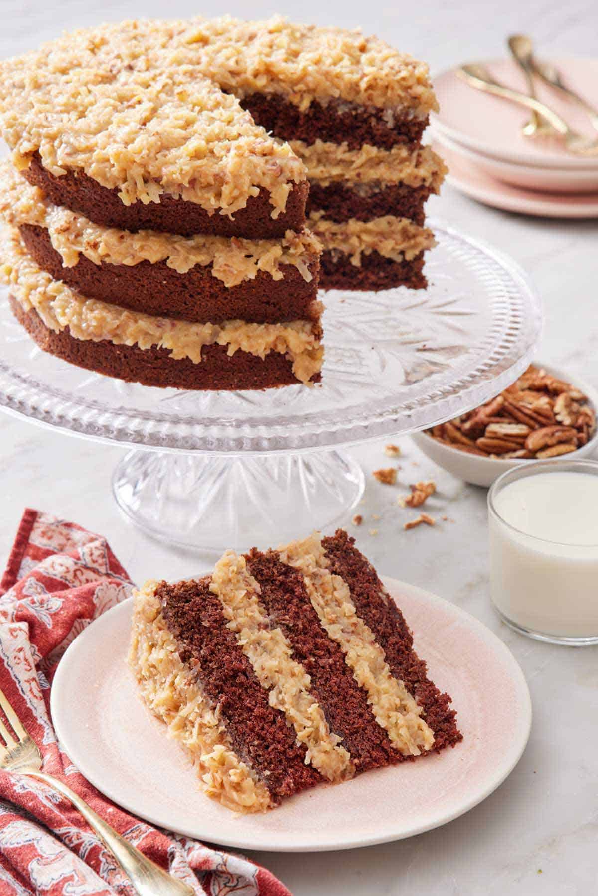 A slice of German chocolate cake on a plate in front of a cake stand with the rest of the cake.