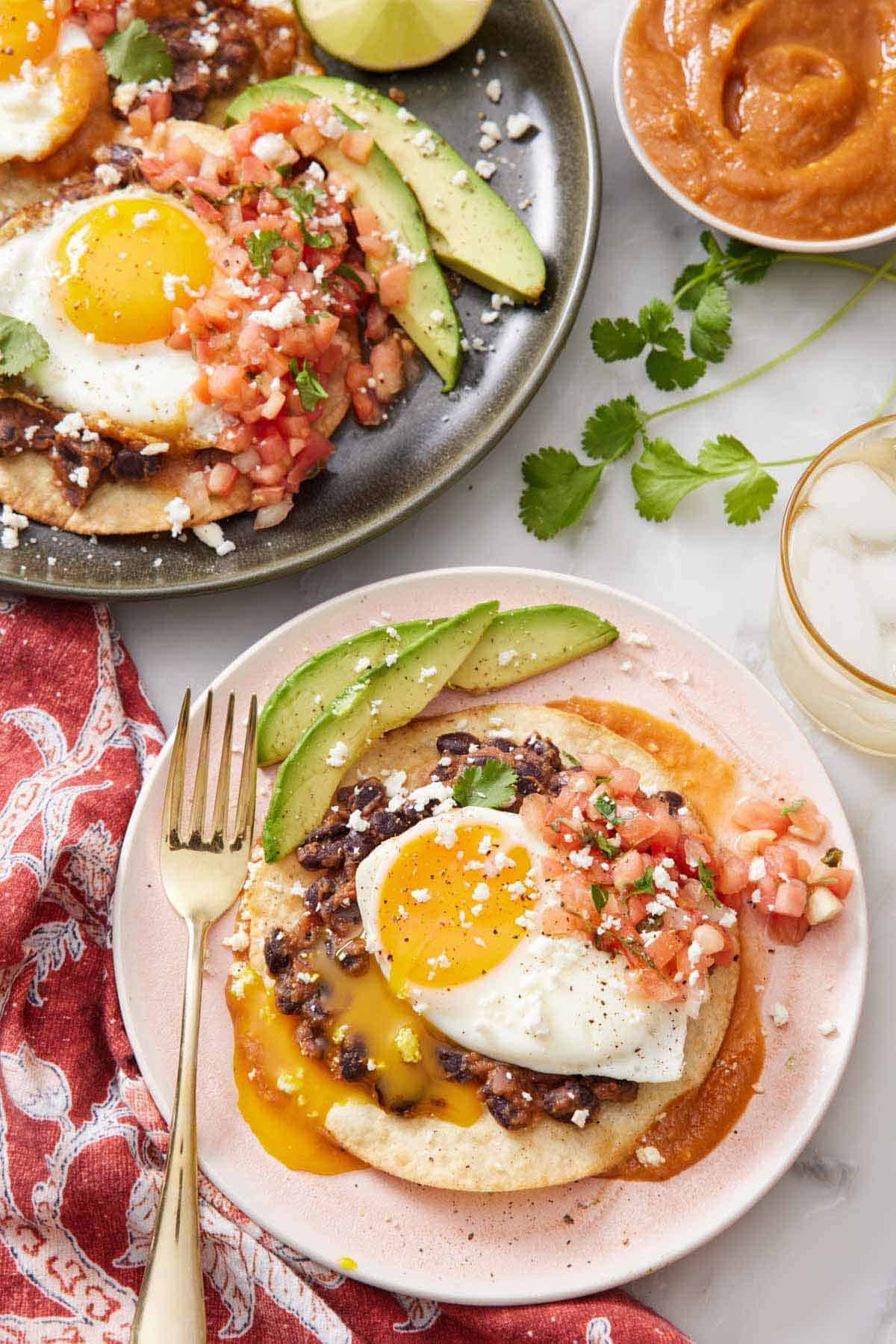 Overhead view of a plate with huevos rancheros topped with cheese, pico de gallo, and sliced avocados. Egg yolk dripping down the dish. Another plate off to the side along with a drink and bowl of sauce.