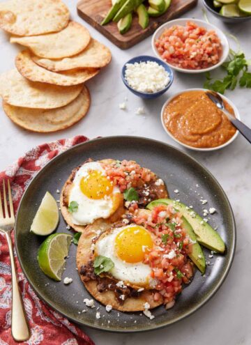 A plate with a serving of huevos rancheros along with lime wedges and avocado slices. Additional toppings in the background and more tortillas.