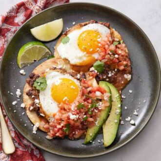 A plate of huevos rancheros with lime wedges, pico de gallo, cheese, and avocado. A fork off to the side.