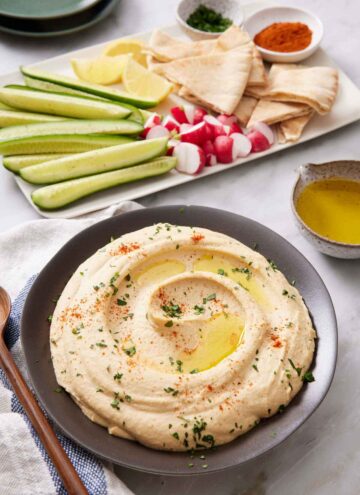 A bowl of hummus topped with oil, paprika, and parsley. Cucumbers, radish, lemons, pita bread, and garnishes in the background on a platter.