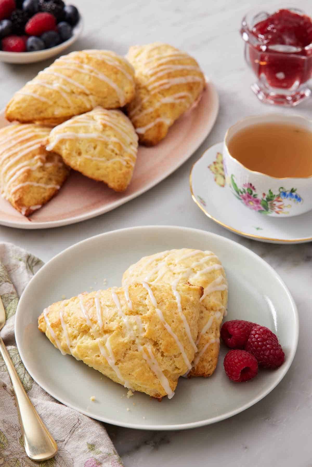 A plate with two lemon scones with fresh raspberries. A cup of tea and additional scones in the background on a platter.