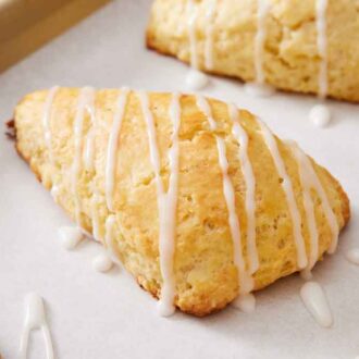 A close up view of lemon scones with glaze drizzled on top on a lined sheet pan.