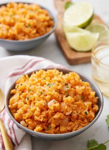 A bowl of Mexican rice with a drink, cut limes, and a second bowl in the background out of focus.