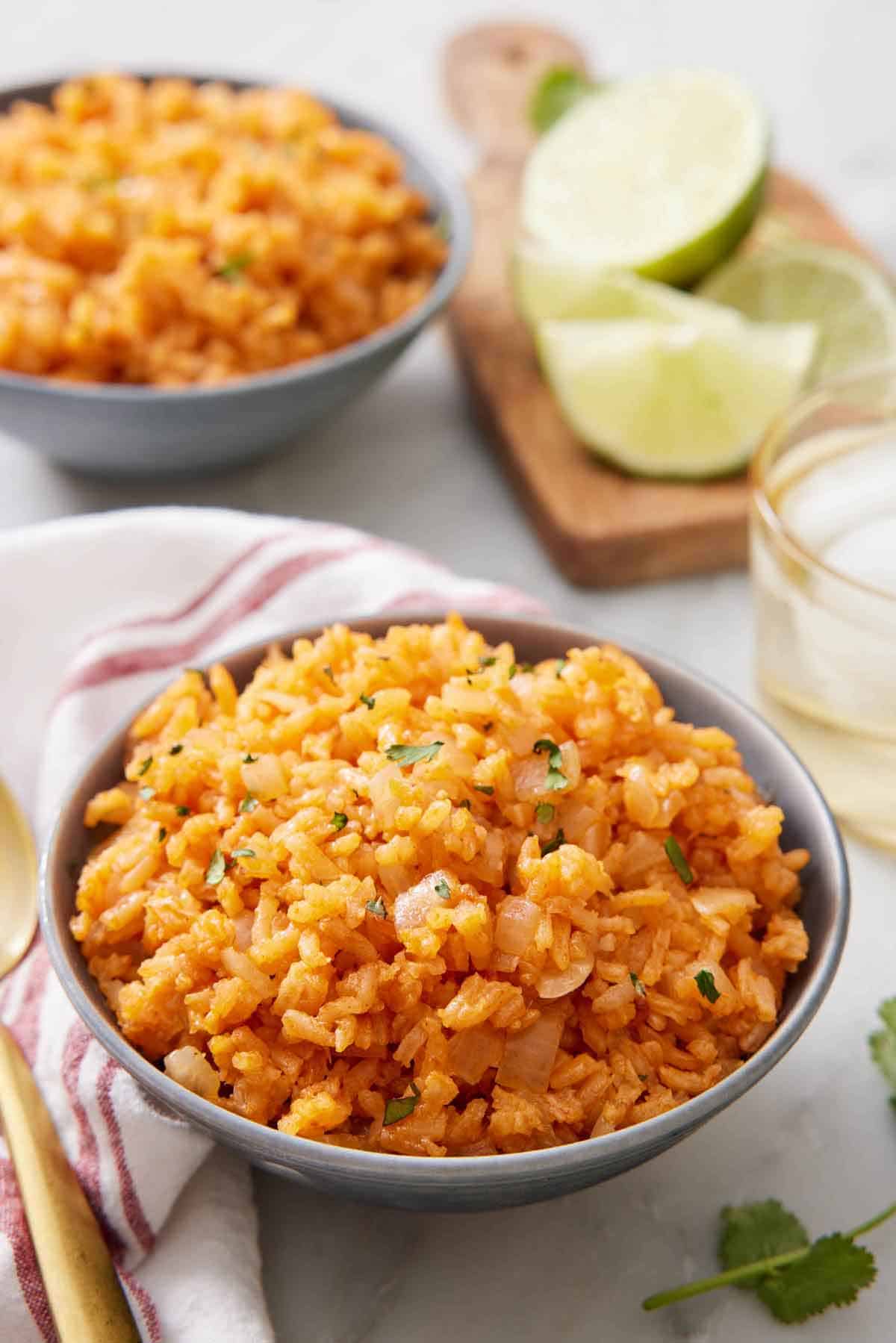 A bowl of Mexican rice with a drink, cut limes, and a second bowl in the background out of focus.