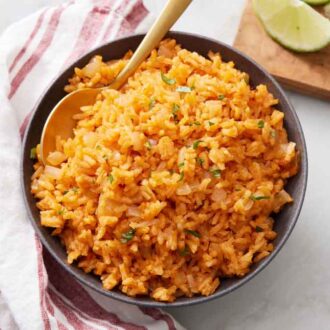 Overhead view of a bowl of Mexican rice with a spoon inside. Cut limes and a napkin on the side.