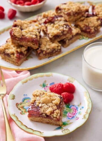 A plate with a piece of raspberry bars with three fresh raspberries. A glass of milk and a platter of more bars in the background.