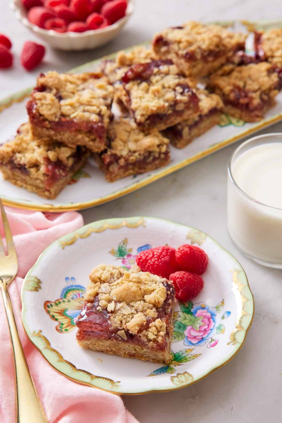 A plate with a piece of raspberry bars with three fresh raspberries. A glass of milk and a platter of more bars in the background.