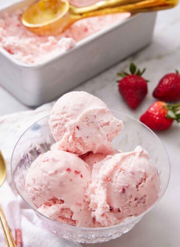 A bowl with scoops of strawberry ice cream with fresh strawberries in the background along with a container of more ice cream.