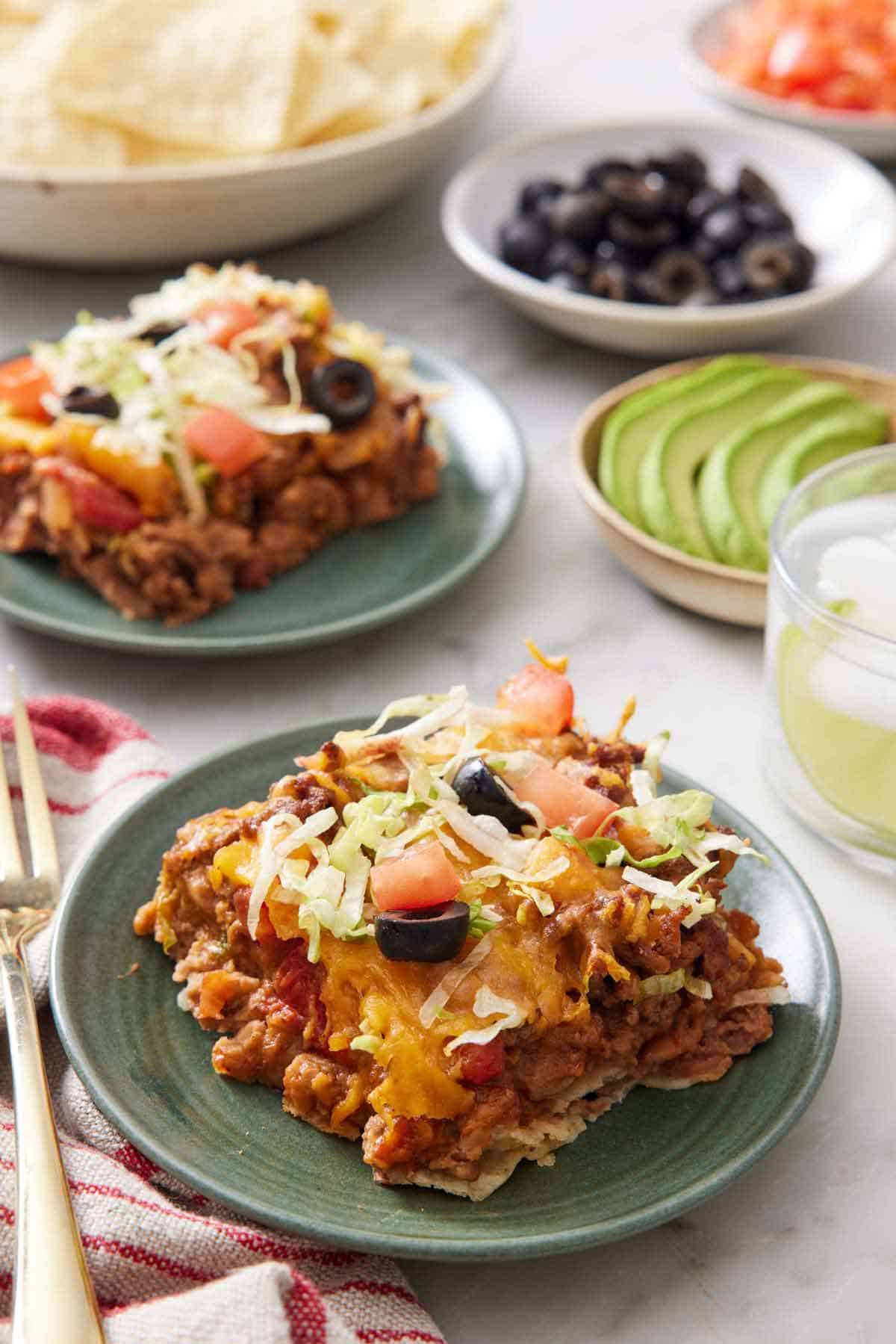 A plate with a serving of taco casserole. Behind it, another plated serving, a drink, a bowl of sliced avocado, a bowl of black olives, and bowl of tortilla chips.